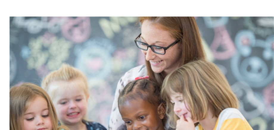 Eine Frau hat zwei Kinder auf dem Schoß, denen Sie etwas aus einem Buch vorliest. Zwei Kinder sitzen neben ihr und schauen mit in das Buch