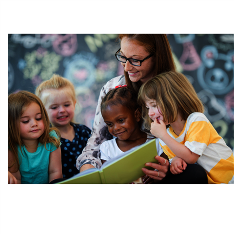 Eine Frau hat zwei Kinder auf dem Schoß, denen Sie etwas aus einem Buch vorliest. Zwei Kinder sitzen neben ihr und schauen mit in das Buch