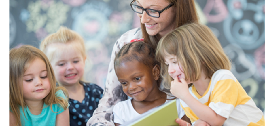 Eine Frau hat zwei Kinder auf dem Schoß, denen Sie etwas aus einem Buch vorliest. Zwei Kinder sitzen neben ihr und schauen mit in das Buch