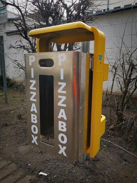 Ein modifizierter Mülleimer aus Edelstahl mit weißer Aufschrift "Pizzabox" auf der Front und Verkehrsgelber Aufhängung auf einem Spielplatz