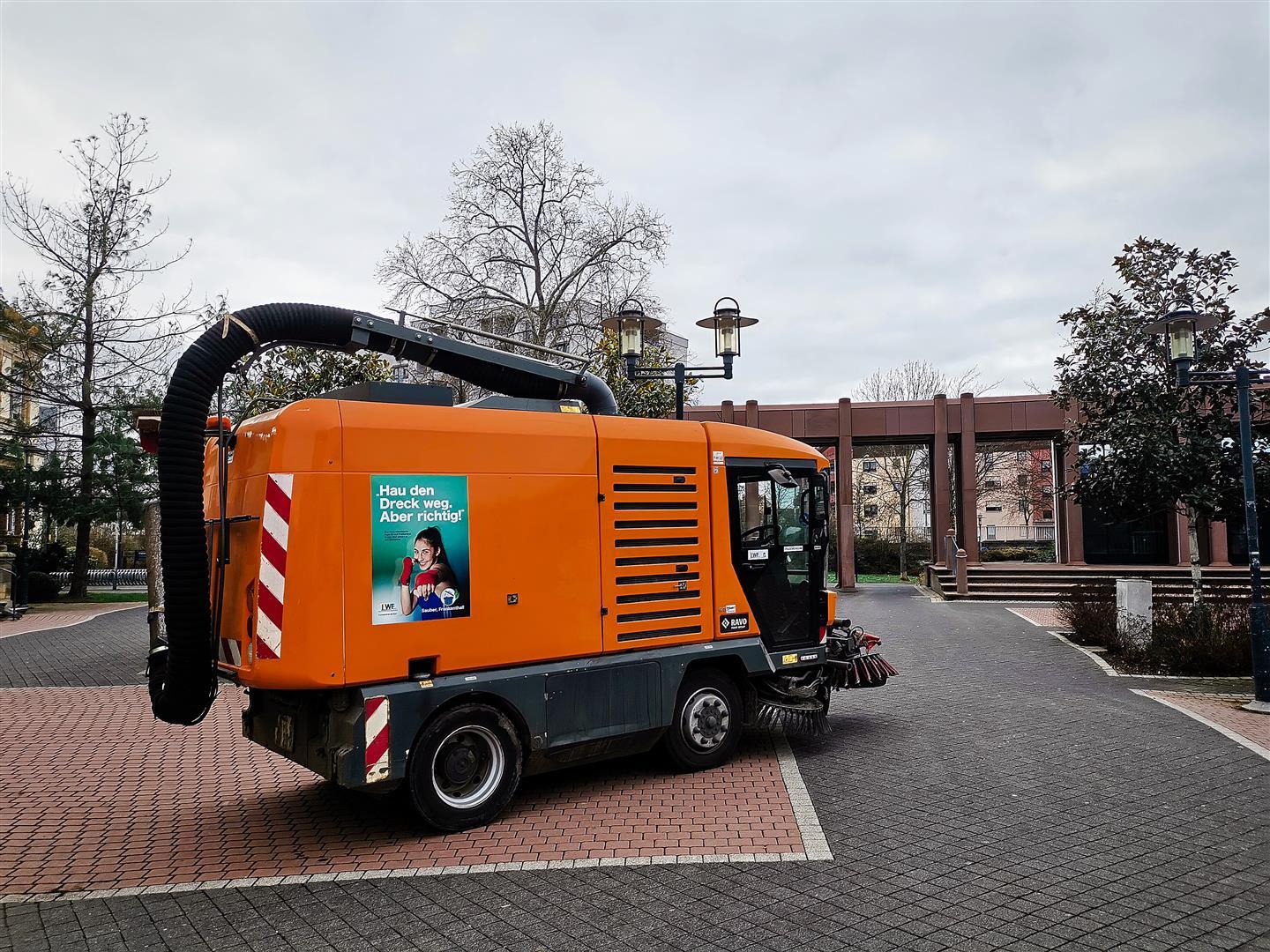 Eine Kehrmaschine mit Saugvorrichtung auf dem Mina Karcher-Platz mit Anti-Littering-Plakat