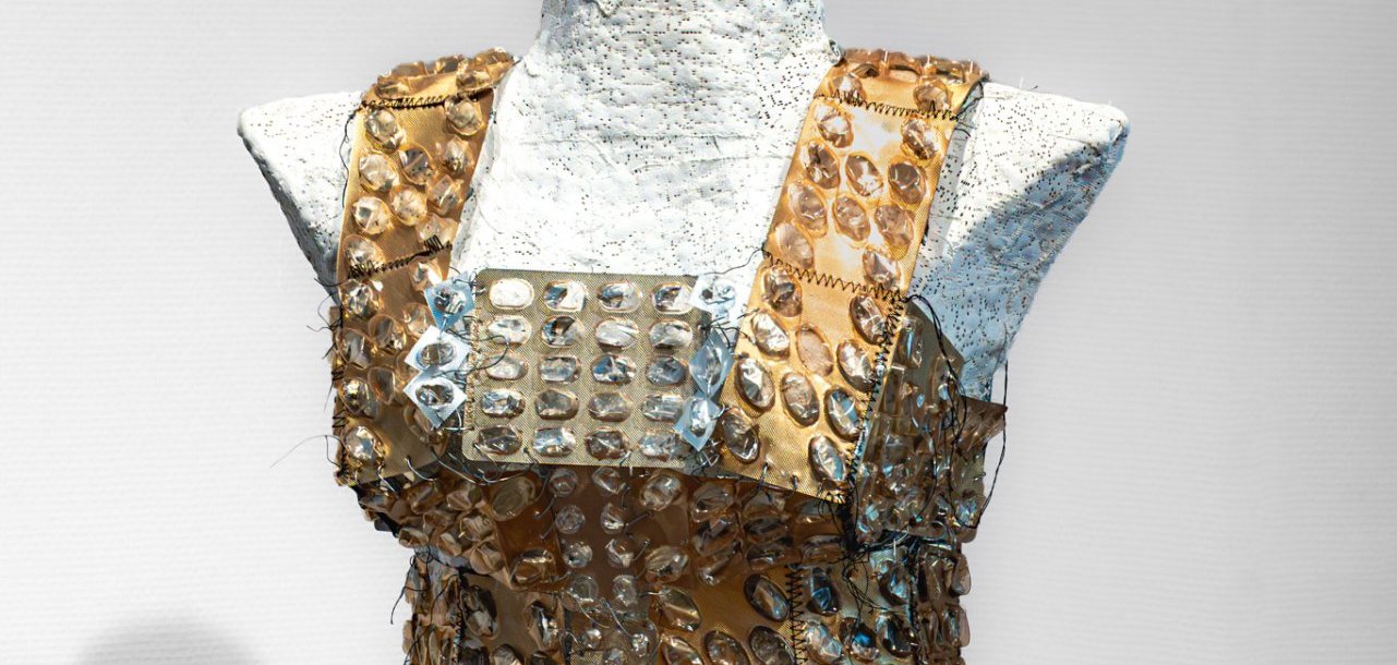 Eine Schneiderpuppe hat ein ärmelloses, beiges Kleid mit transparenten Sticker drauf. Der Hintergrund ist schwarz. Unten steht Freya Blösl, das Leben der Dinge.