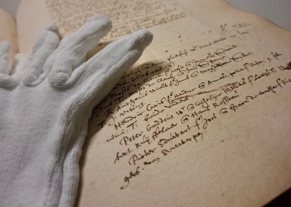 Nahaufnahme eines Eintrags im Kirchenbuch der niederländisch reformierten Gemeinde von 1591, beiliegend ein Baumwollhandschuh