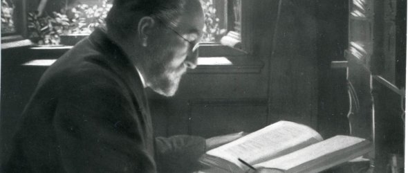 Johann Kraus, Adjunkt der Stadt Frankenthal und Gründer des Frankenthaler Altertumsvereins, sitzt an einem Schreibtisch mit Stift in der einen Hand und einem aufgeschlagenem Buch in der anderen.