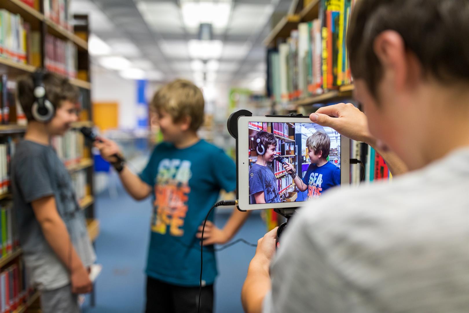 Medien Club Im Vordergrund ist ein Junge der ein Tablet hält in "Kamera Modus". Im Hintergrund sind zwei Jungs. Einer hält ein Mikrofon und der andere hat Kopfhörer auf und spricht in das Mikrofon. im Hintergrund sind Bücherregale