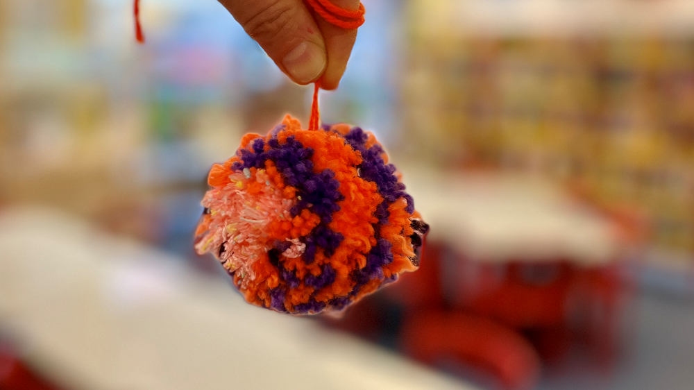 Freitagsaktion Woll-Pompoms basteln Es wird ein Woll-Pompom an einer orangenen Schnurr in die Kamera gehalten. Der Pompom ist orange-lila.