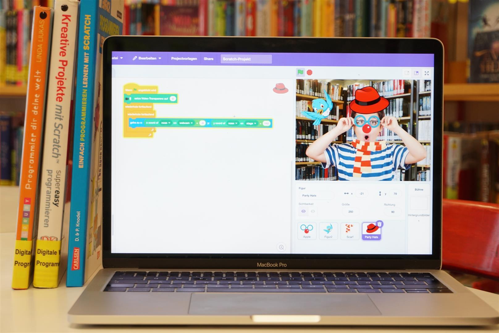 Bild & KI: Gesichtsfilter mit Scratch programmieren Im Hintergrund ist ein Bücherregal zu sehen. Davor steht auf einem Tisch ein aufgeklappter Laptop. In dem Bildschirm sieht man das Programm von Scratch.