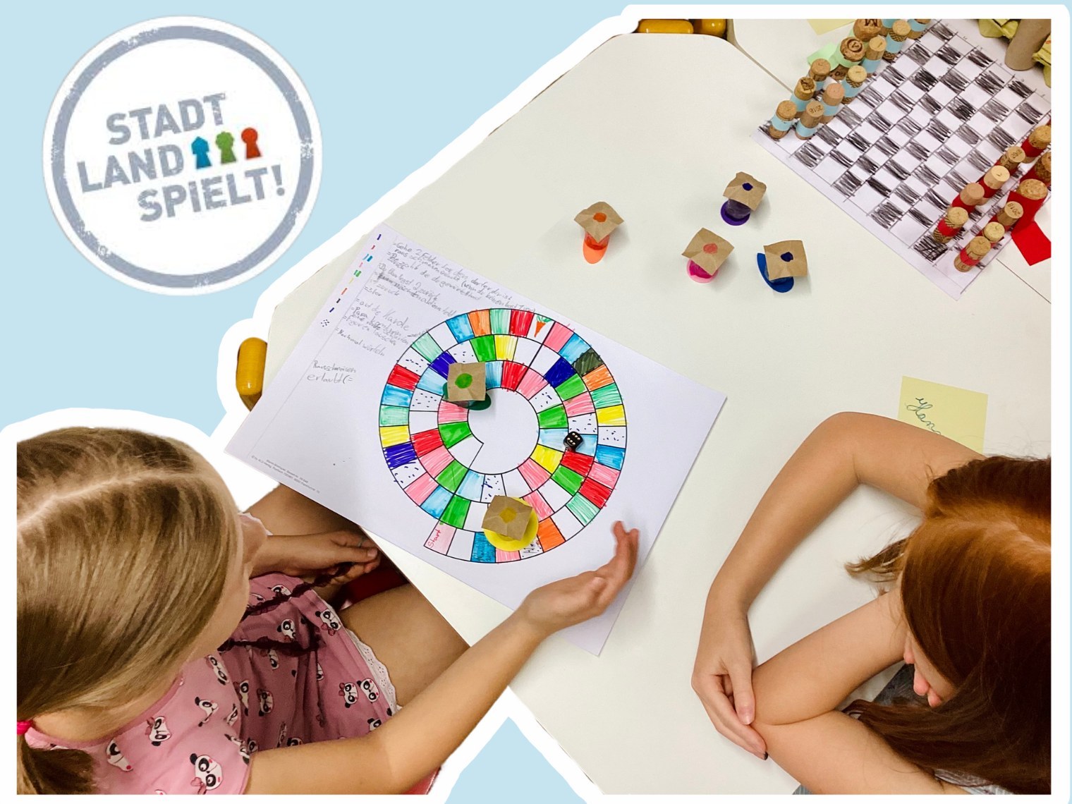 Stadt-Land-Spielt! An einem Tisch spielen 2 Kinder ein Spiel. Oben links in der Ecke ist das Logo von Stadt-Land-Spielt ein weißer Kreis, in dem Stadt-Land-Spielt steht und 3 Spielfiguren in blau, grün rot