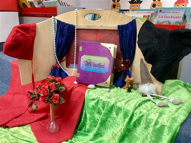 Geschichten für Lesekids auf einer roten und grünen Decke ist das Kamishibai aufgebaut. Im Vordergrund auf den Decken ist eine Vase mit Rosen, ein Reh und ein Buch in Lila mit der Aufschrift Märchen.