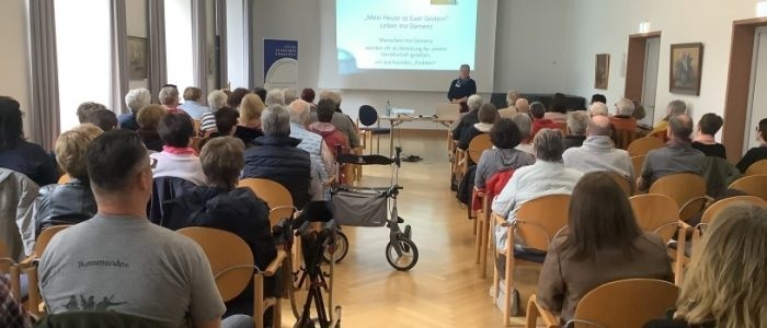 Vortrag Monika Bechtel Demenz Eine Frau steht vor einer Leinwand, auf der "Mein Heute ist euer gestern! ein Leben mit Demenz" zu lesen ist. Davor sitzen viele Menschen auf Stühlen, die ihr zuschauen und -hören, die aber nur mit dem Rücken zu sehen sind