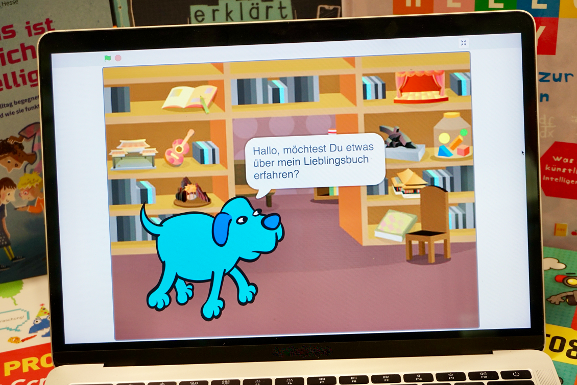 Chatbot programmieren Blick auf einen Laptop-Monitor auf dem im Vollbild ein Chatbot-Programm in der Programmierumgebung Scratch ausgeführt wird. Es ist eine farbige Grafik von einem blauen Hund in einem Zimmer mit Bücherregalen. In einer Sprechblase stellt der Hund die Frage: "Hallo, möchtest Du etwas über mein Lieblingsbuch erfahren?"