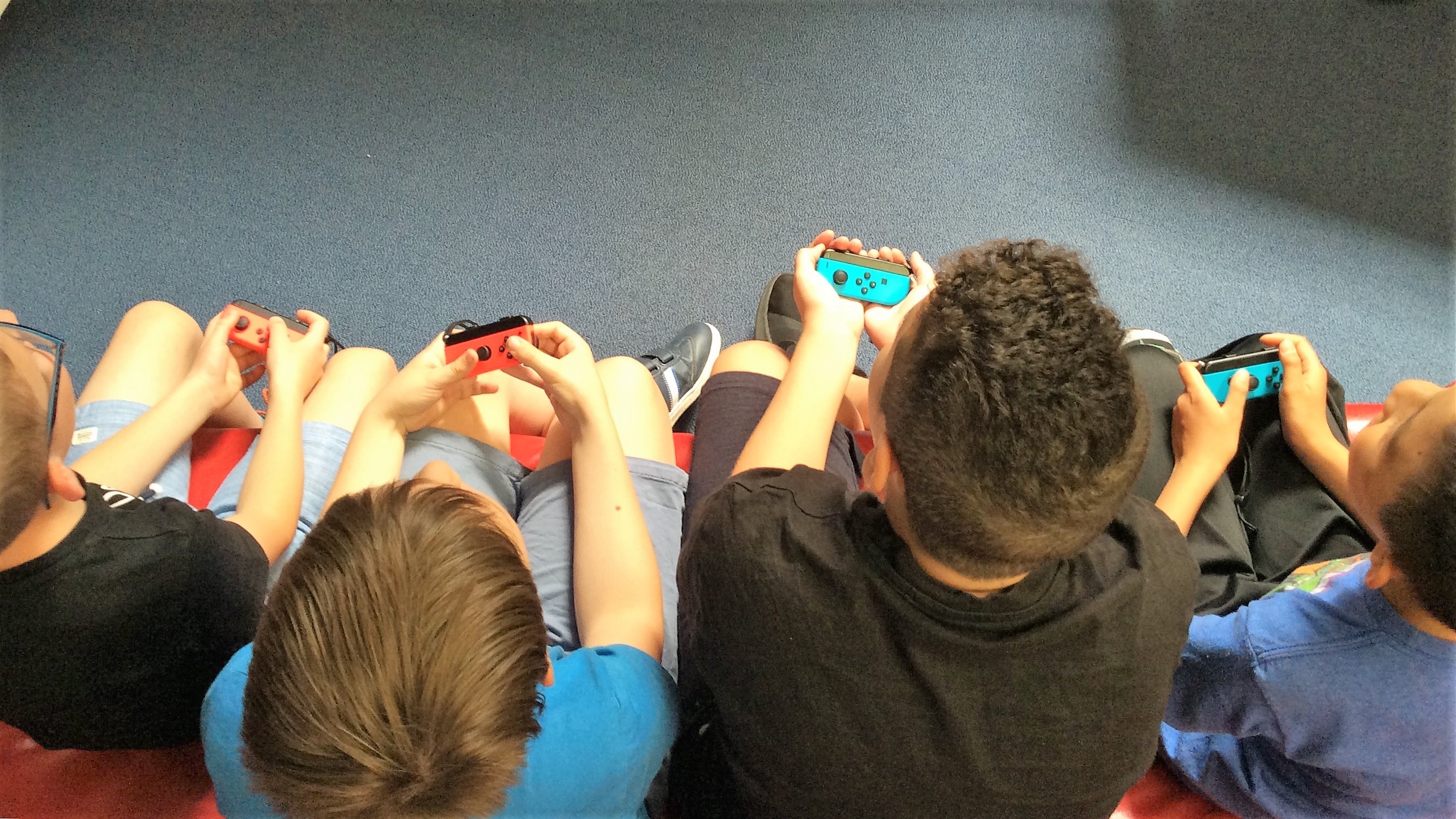 Gaming Club Aus der Vogelperspektive schaut man von hinten auf vier Kinder, die auf einem Sofa sitzen und Switch Controller in den Händen halten.