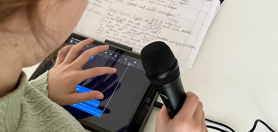 Blick über die rechte Schuler eines Mädchens auf ein Tablet, welches auf einer hellen Tischplatte neben einem handschriftlichen Aufnahmeplan liegt. Mit der linken Hand tippt das Mädchen auf den Bildschirm und bearbeitet ein Audioprojekt. In der rechten Hand hält sie ein Mikrofon.