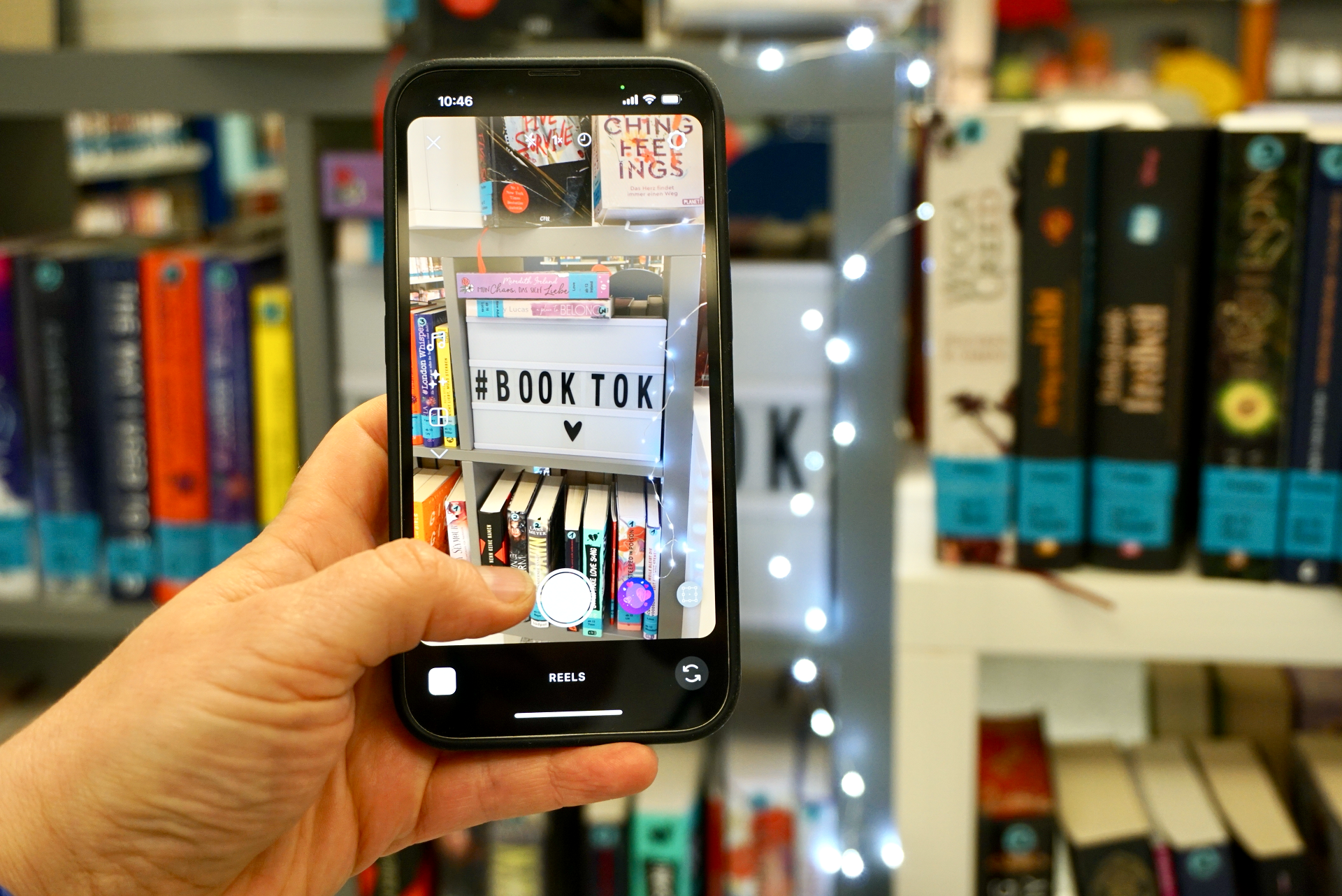Booktok Video-Tricks Im Hintergrund ist in unscharfer Optik ein Bücherregal. Davor wird ein Handy gehalten welches das Bücherregal fotografiert. In dem Bücherregal steht ein Aufsteller in dem Booktok mit einem Herz steht. Um das Bücherregal ist eine Lichterkette