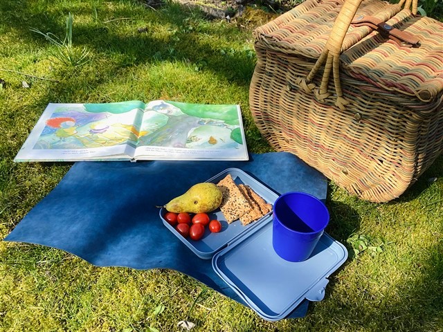 Auf einer blauen Decke liegt ein offenes Bilderbuch, daneben eine Brotdose mit Birne, Kirschtomaten und Knäckebrot sowie ein blauer Becher. Rechts steht ein geflochtener Picknickkorb auf dem Gras in der Sonne.