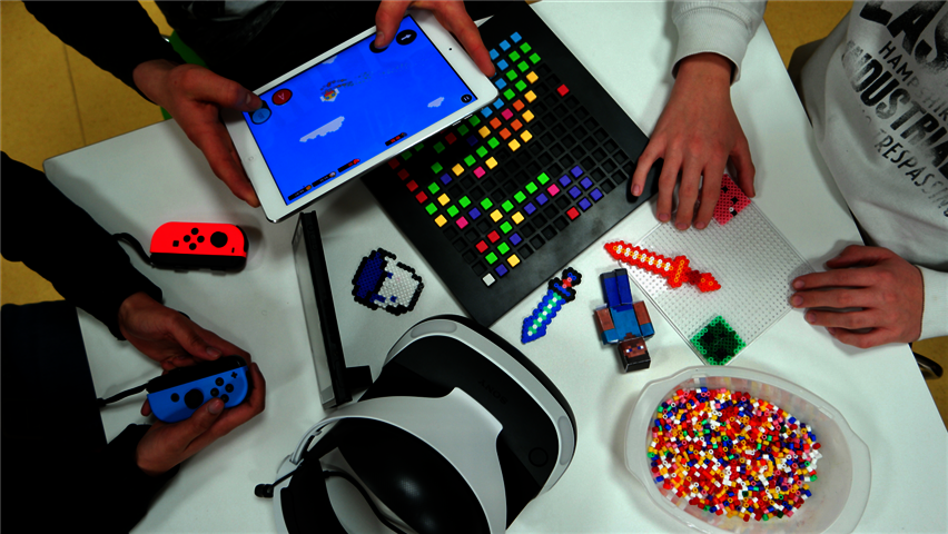 Freitagsaktion - Let´s Play! Gaming zum Kennenlernen, Spielen & Ausprobieren Drei Kinder sitzen an einem Tisch. Ein Kind spielt Nintendo Switch, das andere Kind spielt mit dem IPad Bloxxel und das dritte Kind bastelt mit Bügelperlen