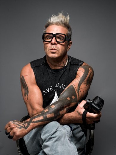 Luigi Toscano Schwarzweiß Foto von einem blonden, a kurzhaarigen Mann mit markanten Tattoos und schwarzem Brillengestell, einen Fotoapparat haltend auf einem Stuhl sitzend.