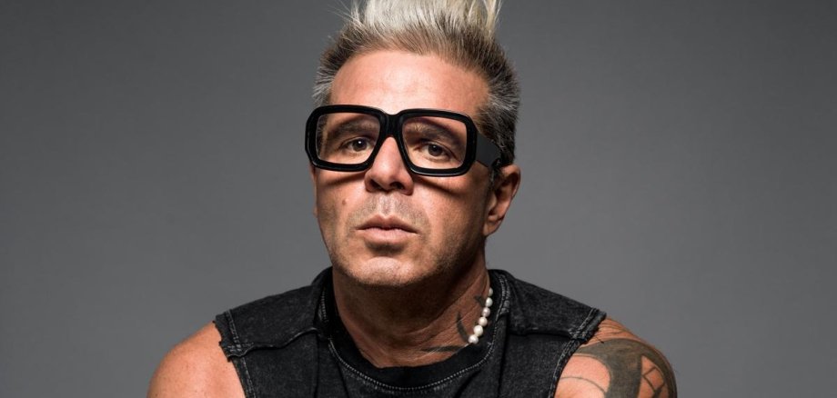 Luigi Toscano Schwarzweiß Foto von einem blonden, a kurzhaarigen Mann mit markanten Tattoos und schwarzem Brillengestell, einen Fotoapparat haltend auf einem Stuhl sitzend.