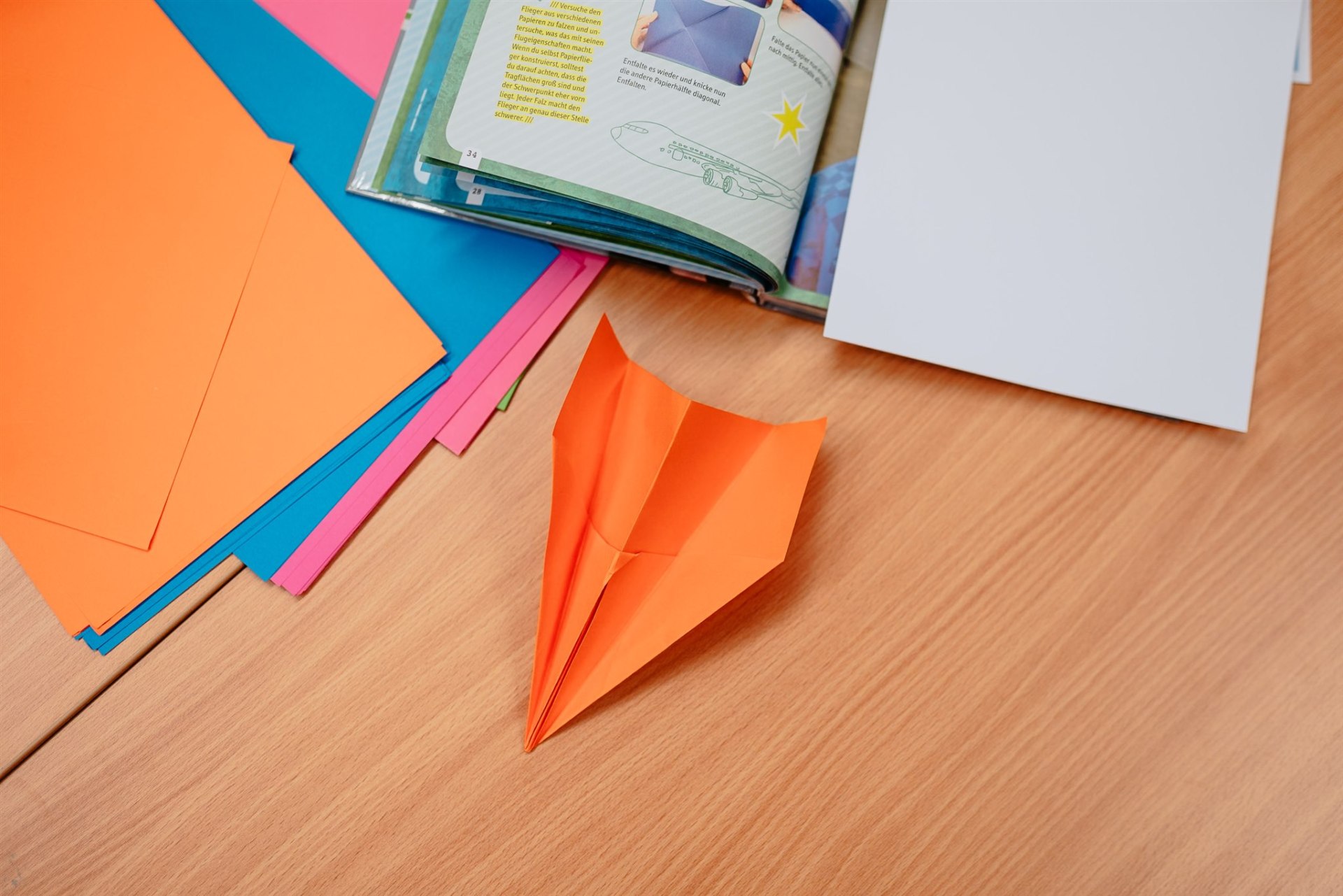 MINTwoch Auf einem Tisch liegen bunte Blätter und ein Papierflieger in orange.