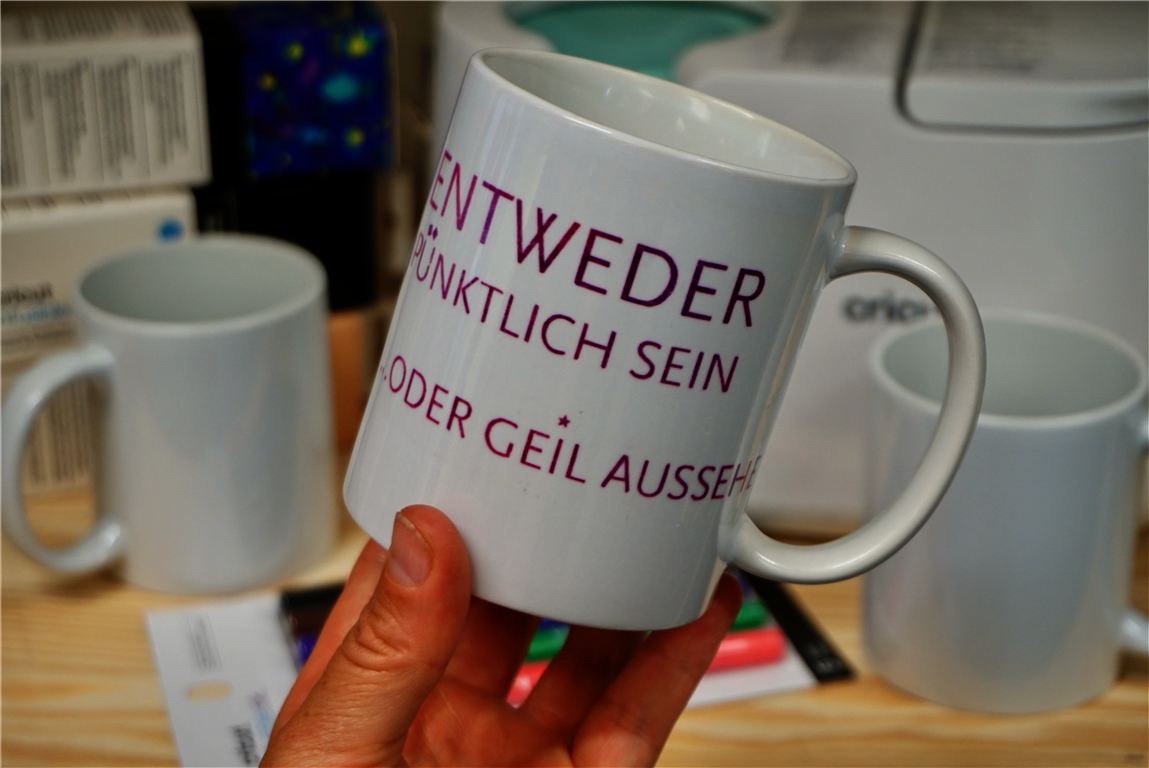 Tassen plotten Eine Hand hält eine weiße Keramiktasse in die Kamera, auf der in violett der Spruch "Entweder pünktlich sein oder gut aussehen" gedruckt ist. Im Hintergrund sind unscharf eine Tassenpresse und Materialien zum Tassen plotten zu erkennen, wie farbige Stifte, weiße Tassenrohlinge und Schachteln mit Plotterfolie.