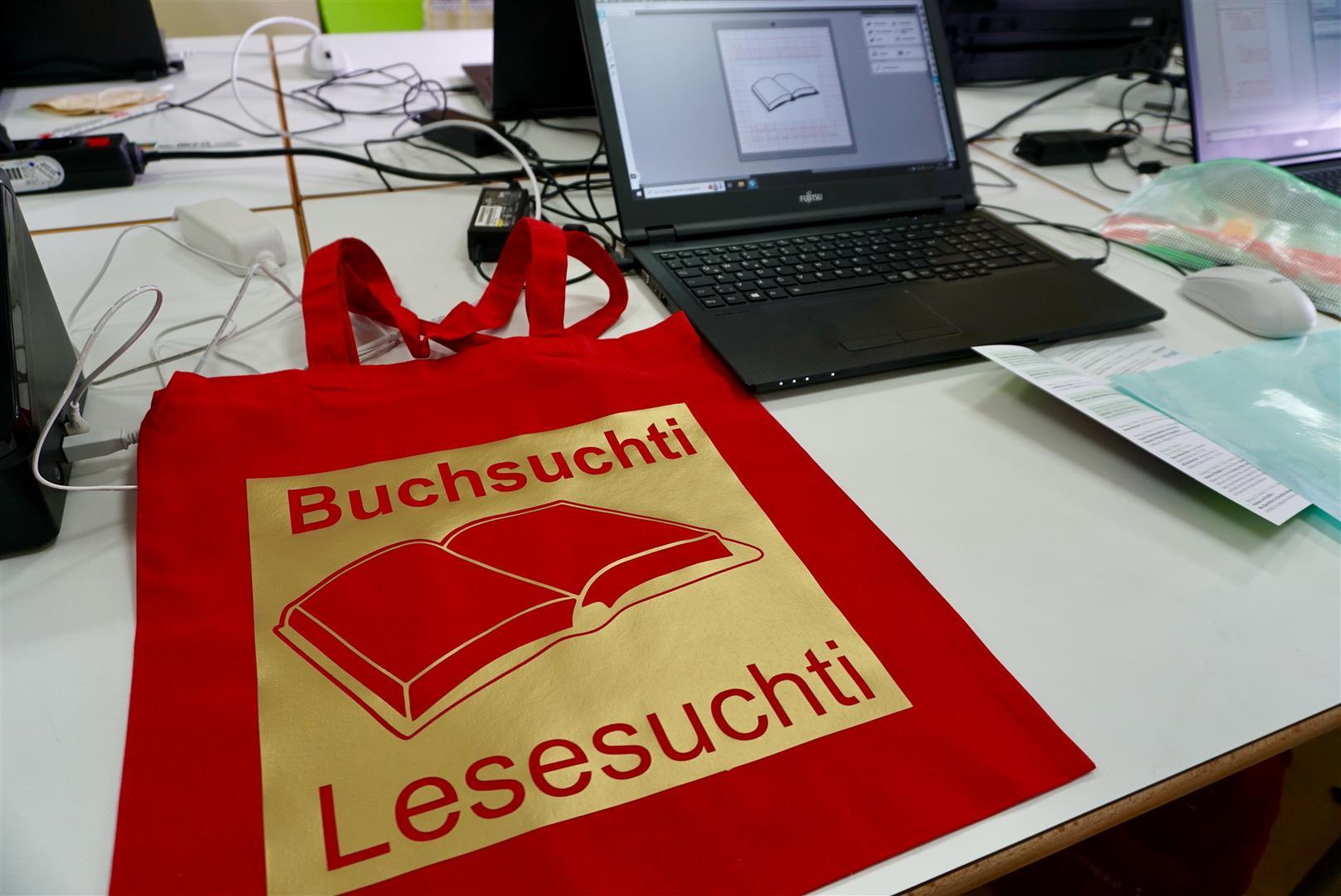 Plotter-Werkstatt Einführung Im Vordergrund liegt auf einem weißen Tisch ein roter Baumwollbeutel, auf dem mit goldener Folie ein Buch und der Schriftzug "Buchsuchti - Lesesuchti" gedruckt ist. Im Hintergrund steht ein aufgeklappter Laptop mit dem Beutel-Design in der Gestaltungssoftware. Mehrere Kabel und weitere Laptops sind teilweise im Bild zu sehen.