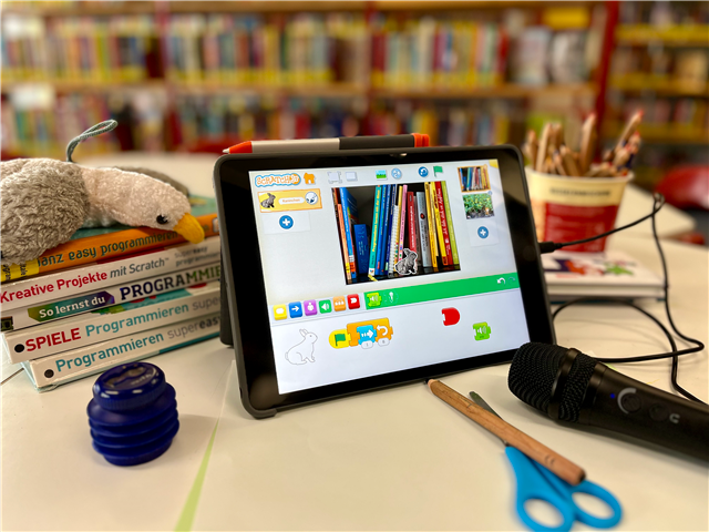 Programmieren mit Scratch Junior Auf einem Tisch in der Kinderbücherei ist ein Tablet aufgestellt, auf dem die App Scratch Junior geöffnet und ein Programm zu sehen ist. Neben dem Tablet liegen außerdem Programmierbücher, Papier, Stifte, Bastelschere und Mikrofon. Im Hintergrund stehen verschwommene Bücherregale.