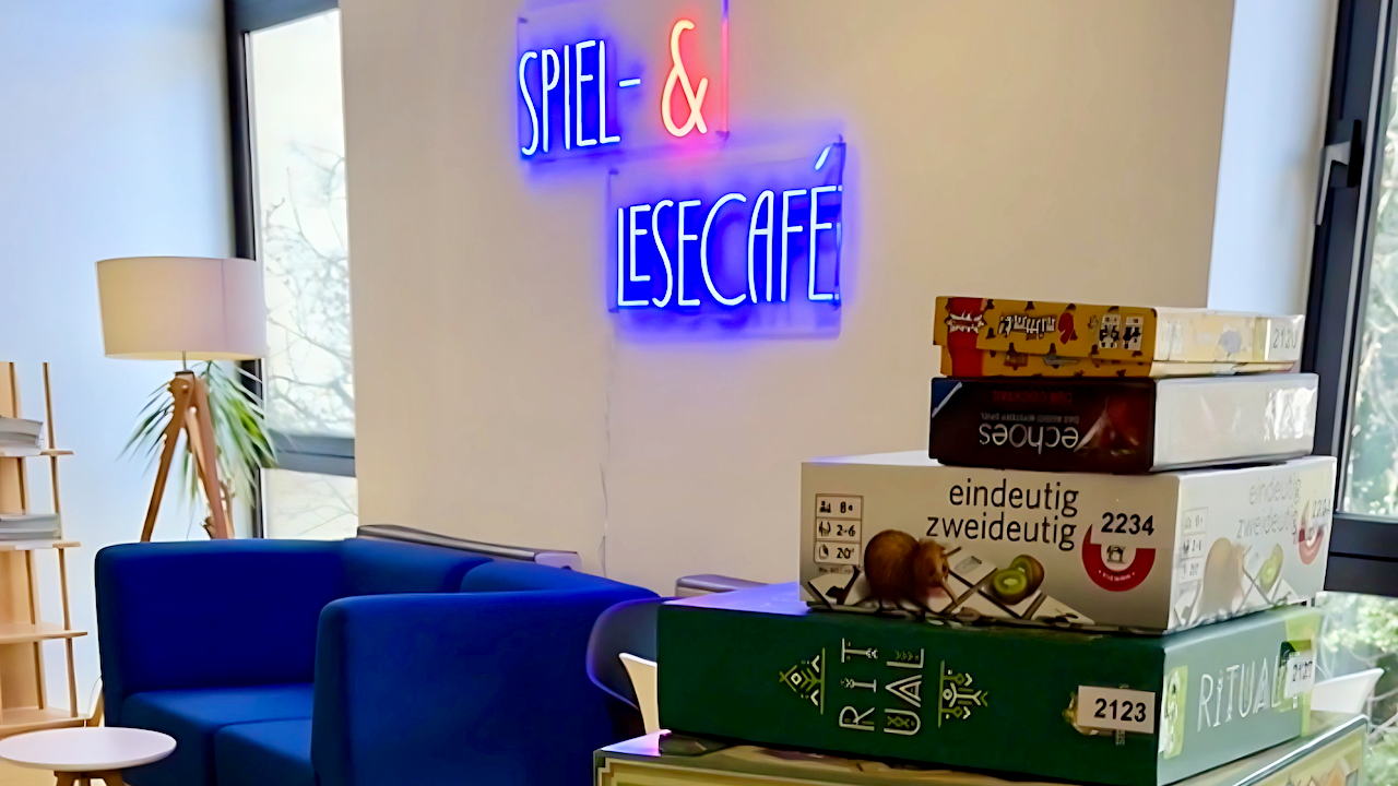 Über einen Stapel von Gesellschaftsspielen im Vordergrund des rechten Bildteils schaut man im Hintergrund auf ein blaues Sofa mit einer weißen Stehlampe auf die blau-orangene Leuchtschrift "Spiel- & Lesecafé", die an einer weißen Wand angebracht ist.