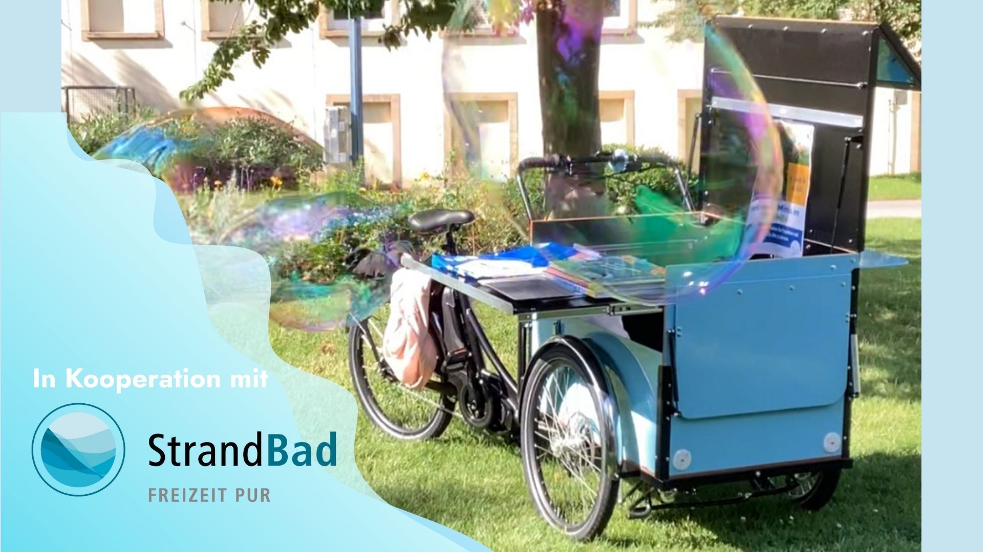 Tag des Freibads In der linken unteren Ecke steht auf hellblauem Hintergrund "In Kooperation mit StrandBad Freizeit pur und das Logo vom StrandBad Frankenthal. Rechts daneben steht auf einer Wiese unter einem Baum ein hellblaues Lastenfahrrad. Davor fliegen zwei große Seifenblasen.