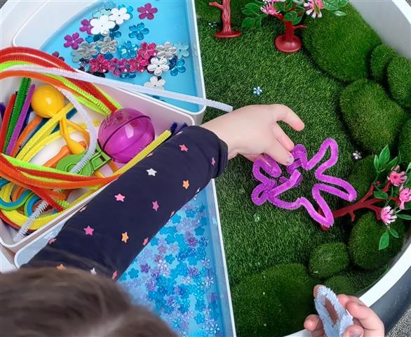 Vorlesen und Erzählen - Clevere Freispiel-Tipps nach Bilderbüchern Blick von oben in eine Schachtel mit Bastelmaterial. Eine Kinderhand setzt einen pinkfarbenen Seifenputzer in Form einer Blume auf grünen Kunstrasen.
