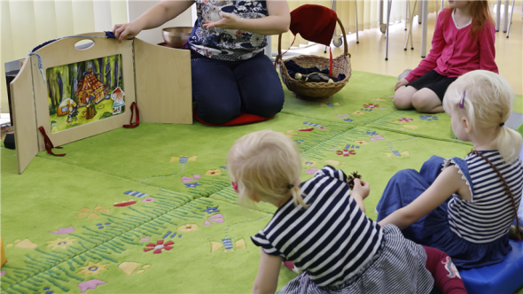 Vorlese Maxis Drei Kinder sitzen auf grünen Teppichen vor dem Kamishibai