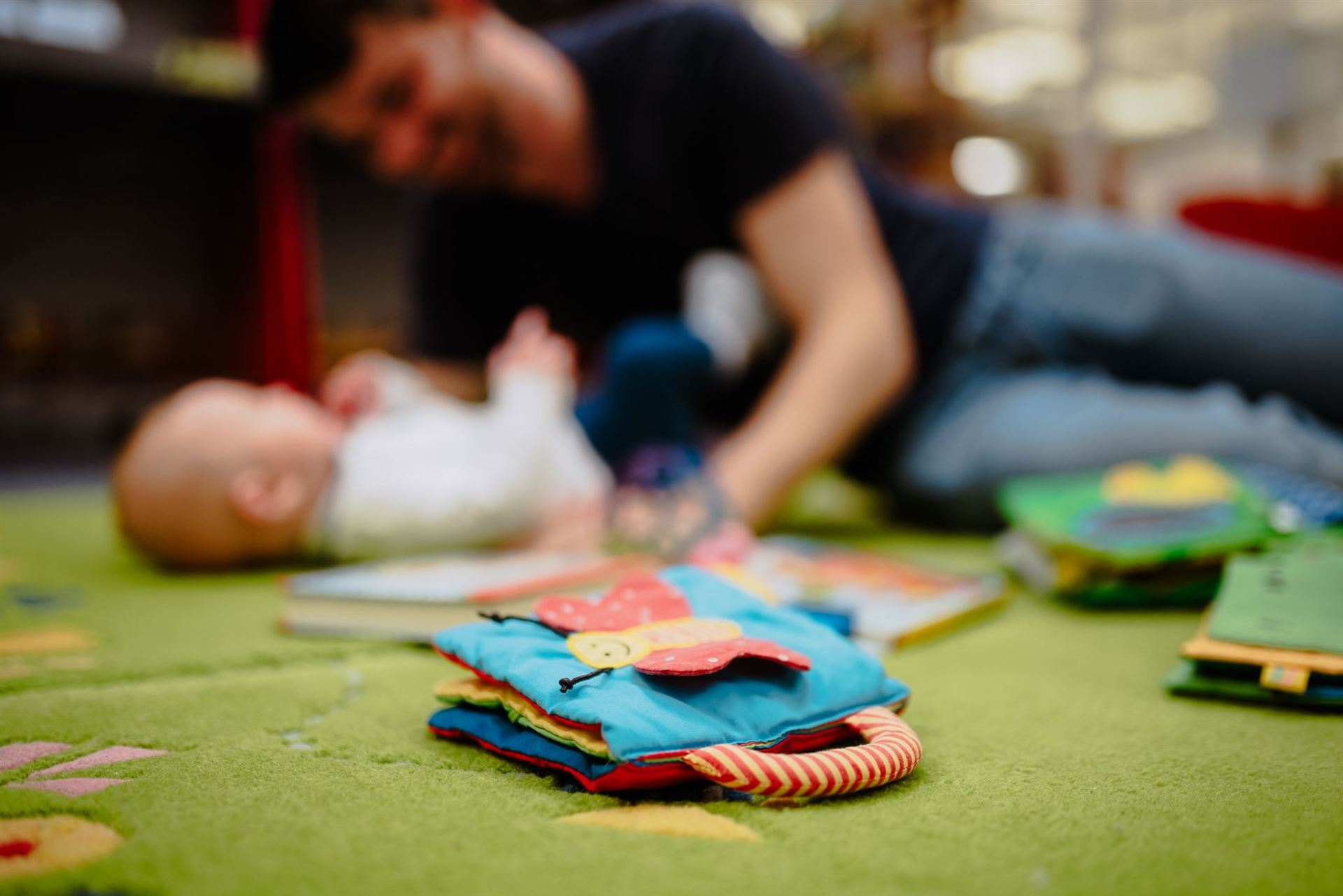 Vorleseminis im Grünen Im Hintergrund unscharf liegt ein Mann mit einem Baby auf einem grünen Teppich. Im Vordergrund scharf ein Stoffbuch für Kleinkinder.
