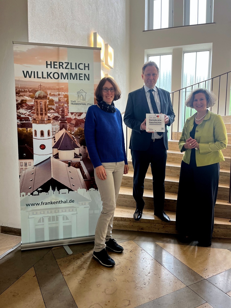 Frankenthal hat die Plakette "Ort der Demokratie-Geschichte" von der Arbeitsgemeinschaft „Orte der Demokratiegeschichte“ in Kooperation mit der „Gesellschaft zur Erforschung der Demokratiegeschichte“ erhalten. Im Bild zu sehen: (von links nach rechts) Stadtarchivarin Dr. Dörte Kaufmann, Oberbürgermeister Martin Hebich und Museumsleiterin Dr. Maria Lucia Weigel im Treppenhaus des Rathauses vor dem Plakat "Herzlich Willkommen. Stadt Frankenthal (Pfalz)". Martin Hebich hält die Plakette.