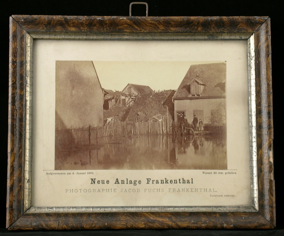 Das Objekt des Monats März 2026: Eine gerahmte Fotografie, die das Rheinhochwasser im Jahr 1882/83 zeigt. In einem Wohnviertel steht Wasser bis zur oberen Treppenstufe eines Hauses, der Zaun ist halb versunken, ein weiteres Haus scheint in sich zusammengestürzt zu sein.