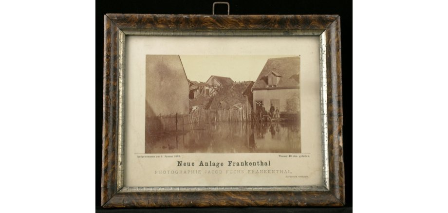 Objekt des Monats März 2026 Das Objekt des Monats März 2026: Eine gerahmte Fotografie, die das Rheinhochwasser im Jahr 1882/83 zeigt. In einem Wohnviertel steht Wasser bis zur oberen Treppenstufe eines Hauses, der Zaun ist halb versunken, ein weiteres Haus scheint in sich zusammengestürzt zu sein.