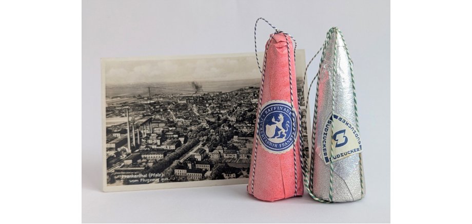 Objekt des Monats April 2026. Zu sehen: Eine Schwarz-Weiß-Ansichtskarte mit einer Luftbildaufnahme der Zuckerfabrik mit ihren sieben Schornsteinen vor der Zerstörung im Zweiten Weltkrieg, davor zwei originalverpackte Zuckerhüte aus der Zeit der Zuckerfabrik. Ein Zuckerhut ist zartrosa verpackt mit Etikett. Mittig auf dem Etikett ist der Frankenthaler Löwe mit dem Frankenthaler Wappen, silbern vor blauem Hintergrund, und der in den gleichen Farben gehaltenen Umschrift "Raffinade Zuckerfabrik Frankenthal". Der andere, in silberner Glanzverpackung eingehüllte Zuckerhut trägt ein mit mittig dem Logo der Südzucker blau auf weißem Hintergrund und als Umschrift 3x das Wort "Südzucker" ebenfalls blau auf weißem Hintergrund. Beide Zuckerhüte sind zusätzlich mit einer blau-weißen Kordel verpackt.