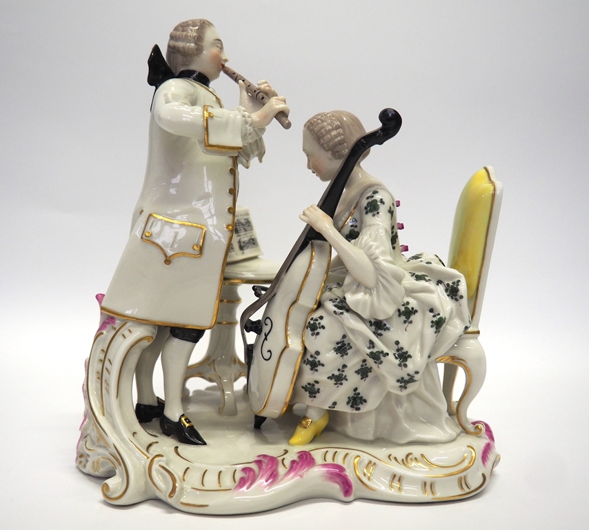 Allegorie "Das Gehör" Nymphenburger Porzellangruppe: Allegorie "Das Gehör". Auf einem gelb gepolsterten Lehnstuhl sitzt eine Cello spielende Frau, die auf dem Notenständer auf dem vor ihr stehenden ovalen Tisch blickt. Sie trägt ein grün geblümtes Kleid mit Volants und rosa aufgesetzten Schleifen an der Schnürung im Rücken. Auf dem Kopf sitzt eine gelockte Perücke. Ihr gegenüber steht ein Herr im weißen Justaucorps über der weißen Weste, mit Beutelperücke und schwarzer Kniebundhose, der eine Querflöte bläst. Als Rückenstütze dient dem Mann ein flacher Rocaillewedel. Die Gruppe steht auf einem flachen, von rosa Rocaillen umsäumten Sockel.