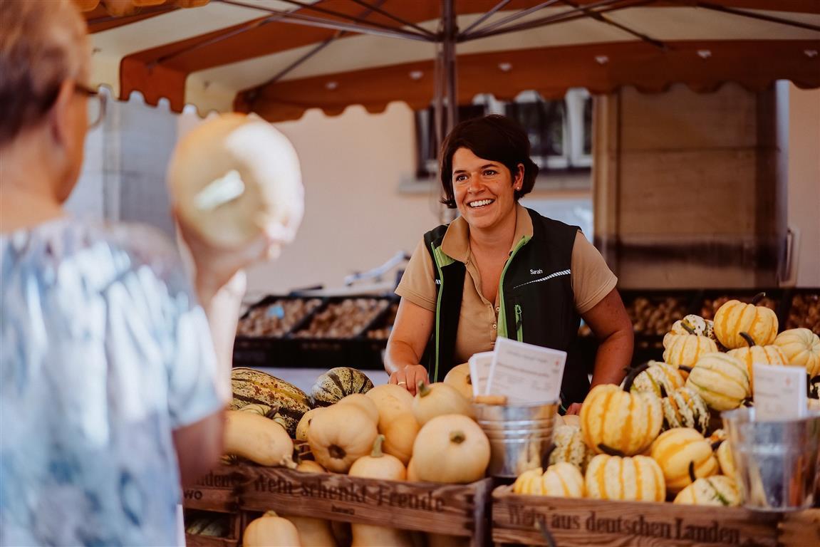 Marktstand vom Erbeer- und Spargelhof Schreiber. Frau Schreiber breit lächelnd zwischen Kürbissen. Im Vordergrund eine weitere Frau.