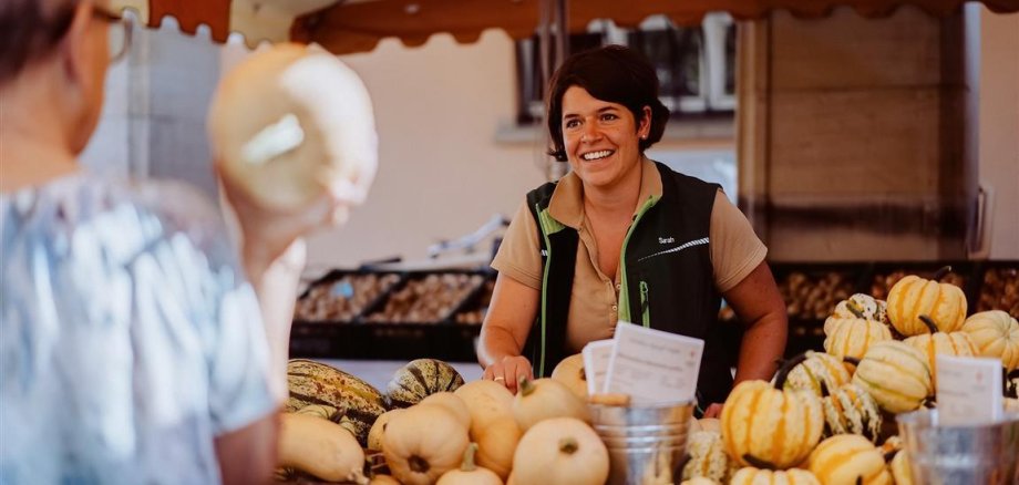 Marktstand vom Erbeer- und Spargelhof Schreiber. Frau Schreiber breit lächelnd zwischen Kürbissen. Im Vordergrund eine weitere Frau.