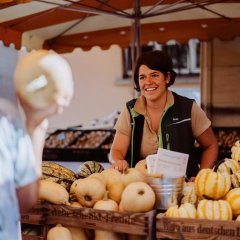 Marktstand vom Erbeer- und Spargelhof Schreiber. Frau Schreiber breit lächelnd zwischen Kürbissen. Im Vordergrund eine weitere Frau.