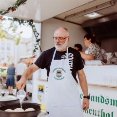 Marktstand der Donaudeutsche Landsmannschaft Frankenthal e.V., ein Mann brät Dampfnudeln in einer großen Pfanne. Im Hintergrund Frauen in einem Anhänger.