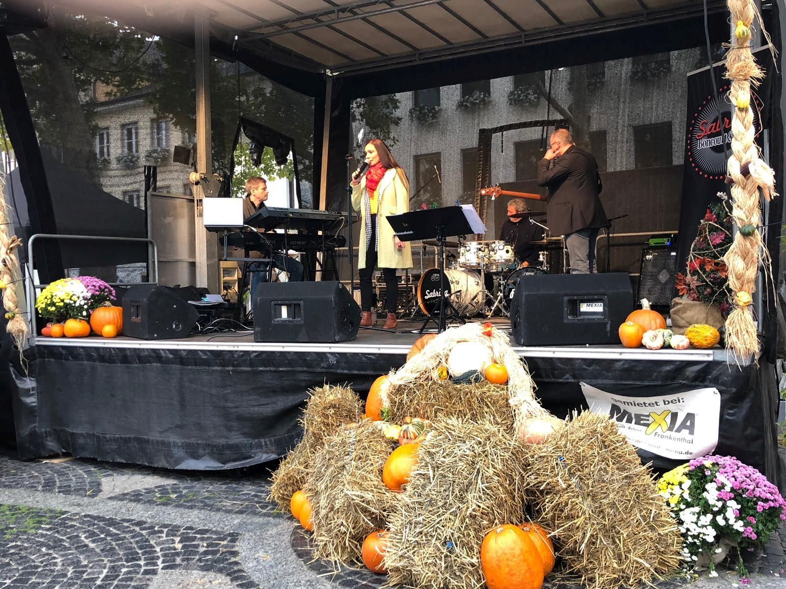 Vierköpfige Band aus einer Sängerin und drei Musikern auf einer Bühne. Diese ist herbstlich dekoriert.