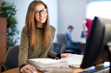 Junge Frau sitzt an einem Schreibtisch mit PC und lächelt.
