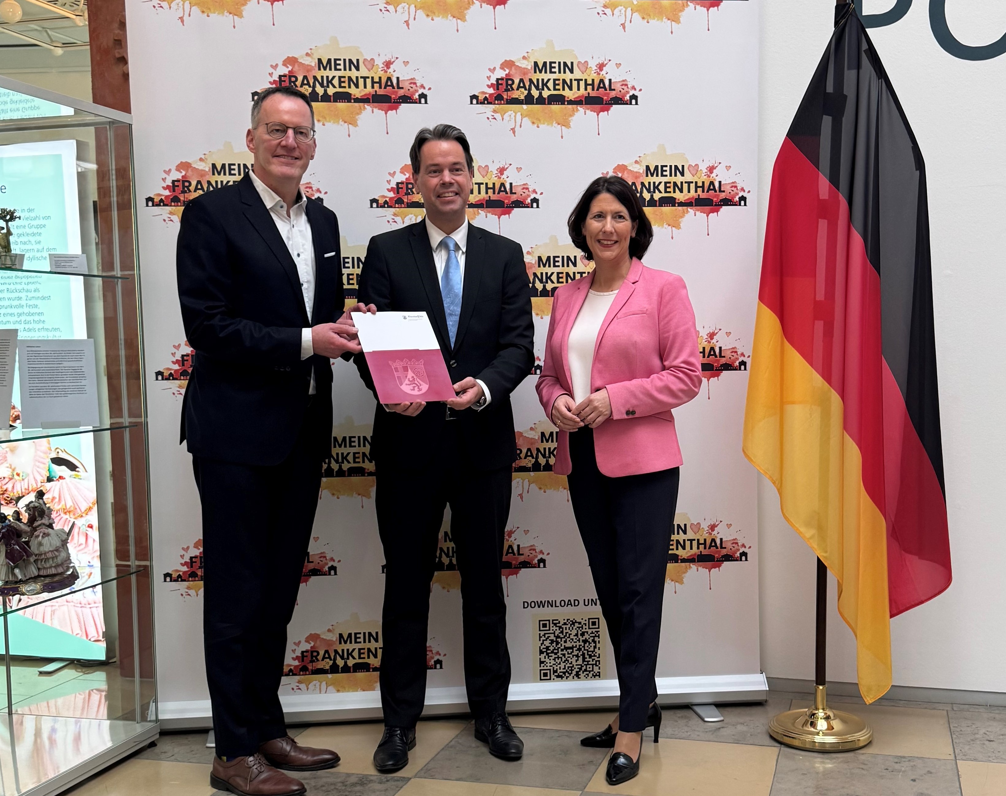 Zwei Herren im Anzug und eine Frau stehen neben einer Deutschlandfahne. Die Herren halten gemeinsam ein Dokument. 