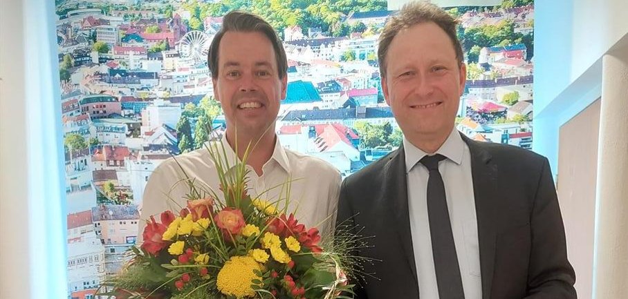 Zwei lächelnde Männer, von denen einer einen Blumenstrauß hält.