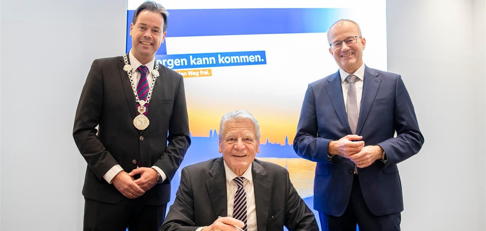 3 Herren in Anzügen, der mittlere schreibt in ein Buch.