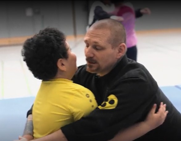 Inklusiver Selbstverteidigungskurs für Menschen mit und ohne Behinderung Herr Beyer im schwarzen Kampfanzug hält einen Jungen im gelben Tshirt fest.
