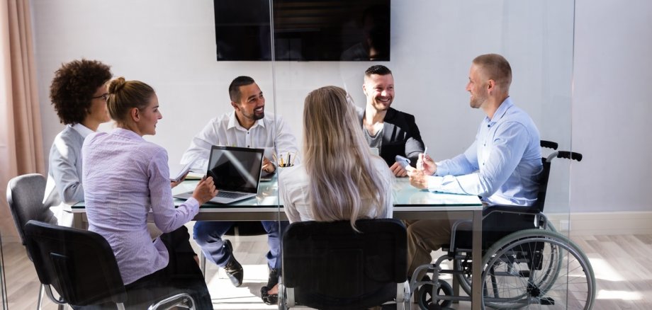 Fünf Menschen und eine Person im Rollstuhl sitzen um einen Tisch herum und besprechen. 