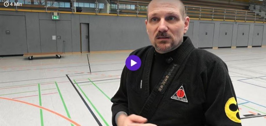 Herr Beyer steht in der Halle und schaut in Richtung Kamera und hält beide Hände vor seinem Körper. Er trägt schwarzen Ju Jutsu Anzug. 