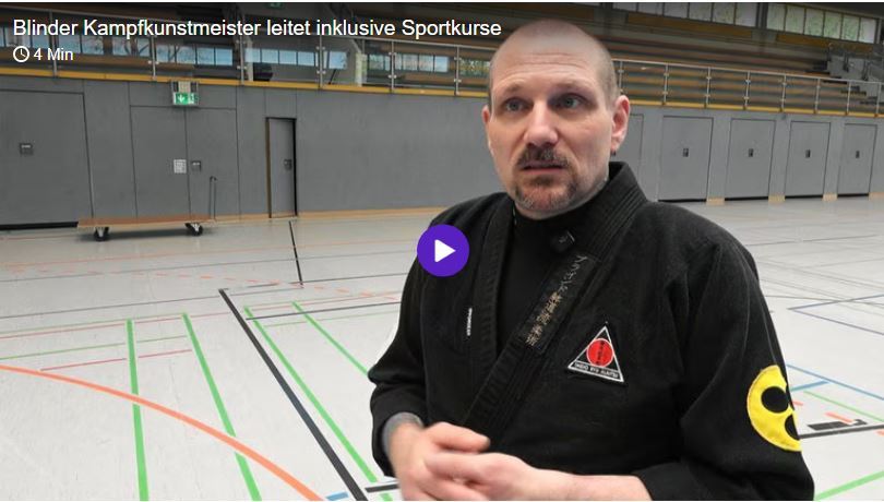 Nachbericht: Blinder Kampfkunstmeister leitet inklusive Sportkurse Herr Beyer steht in der Halle und schaut in Richtung Kamera und hält beide Hände vor seinem Körper. Er trägt schwarzen Ju Jutsu Anzug.