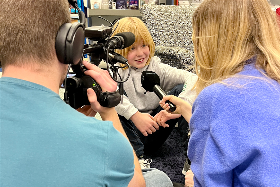 Blick über die Schultern eines Kameramannes und einer Reporterin auf ein Mädchen, welches in ein Mikrofon spricht und von einer Videokamera gefilmt wird. Alle sitzen auf dem Boden in der Stadtbücherei.